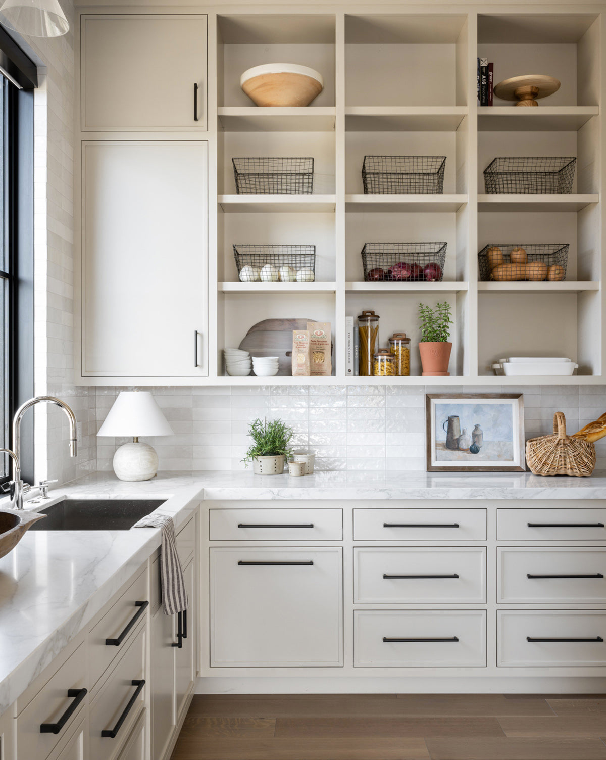 Bright modern kitchen with white cabinets, open shelves displaying baskets, bowls, and Gathered Jars—a stylish mix of home decor and function—enhanced by a marble countertop, framed picture, potted plants, and abundant natural light.