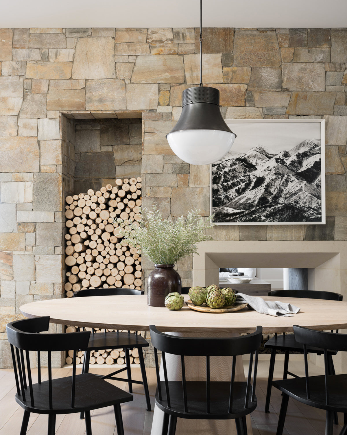 A modern dining area with a round wooden table, Reeves Dining Chairs in black, a vase of greenery, stacked firewood on a stone wall, a mountain landscape photo, and a black pendant light overhead, by McGee & Co.