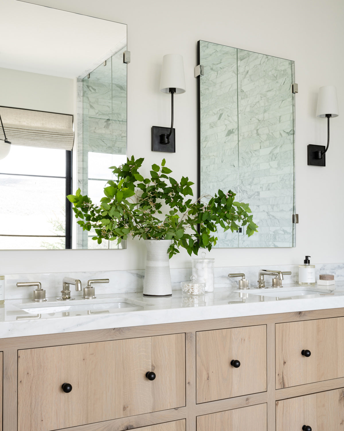 Modern bathroom featuring a light wood double vanity, white marble countertop, two Ivana Mirrors, burnished brass wall sconces, a vase with green branches, and window reflections with a roman shade, by McGee & Co.