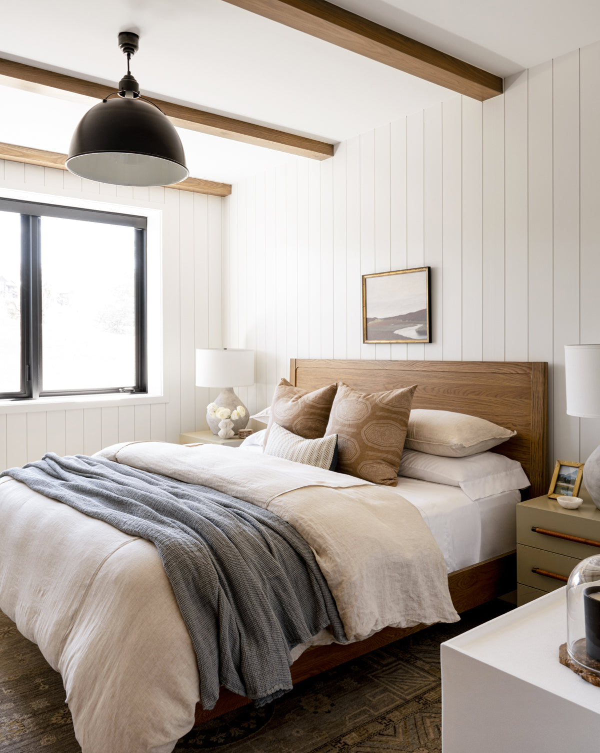 Modern bedroom with white shiplap walls, wooden beams on the ceiling, a wooden bed with beige and gray bedding, two nightstands topped with elegant Eugene Pendant lights, a window, and a small landscape painting above the bed, by McGee & Co.