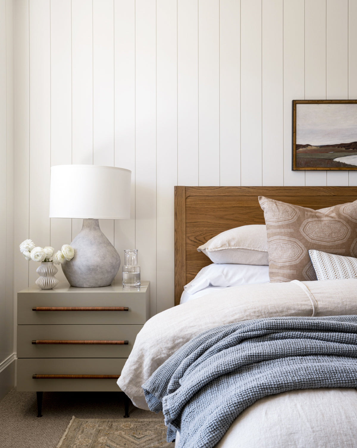 A cozy bedroom features a wooden headboard, neutral bedding with a blue textured blanket, and a nightstand topped with the Stetson Table Lamp, set against white shiplap walls adorned with landscape art above, by McGee & Co.