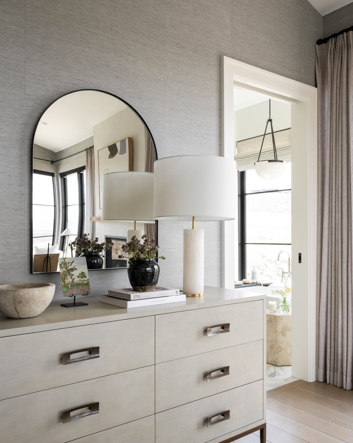 A modern dresser holds a white lamp, books, a bowl, and a vase with flowers. The Wylie Arched Mirror with shatterproof glass reflects windows and wall art as sunlight streams in through an open doorway and curtained window, by McGee & Co.