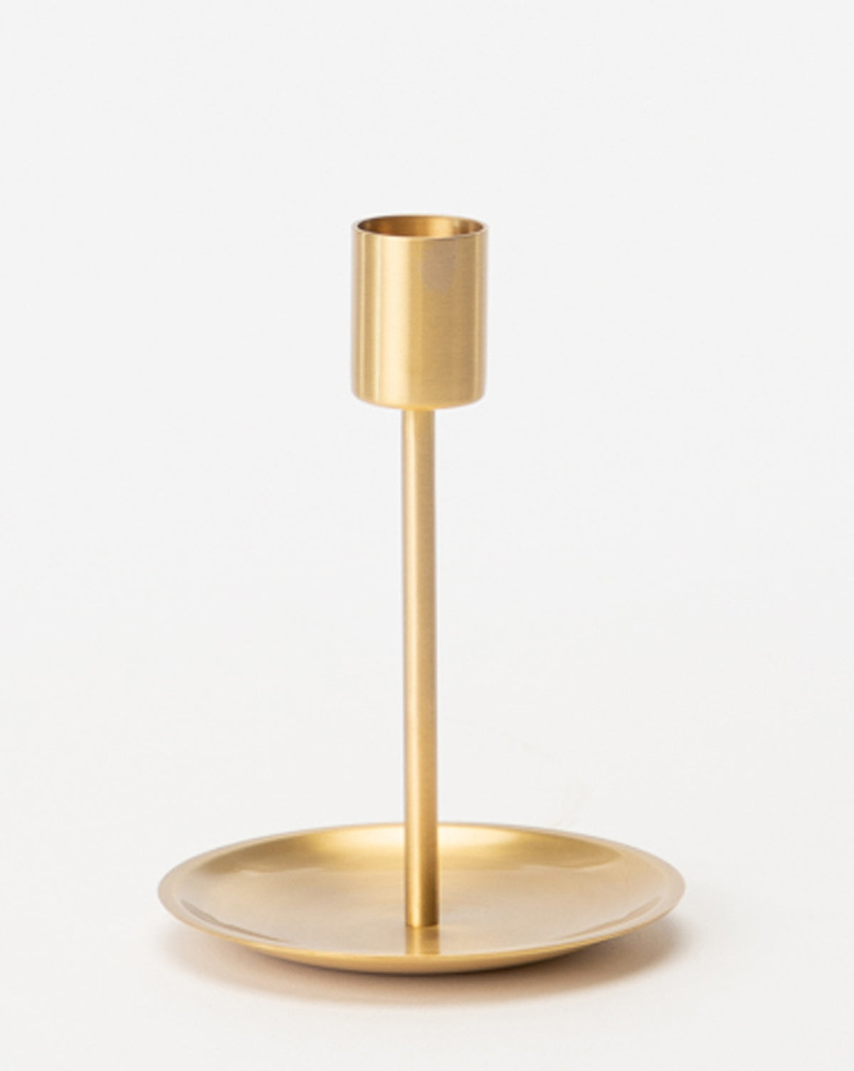 The Brass Taper Holder features a minimalist design with a cylindrical socket, slim stem, and round base, displayed on a plain white background.