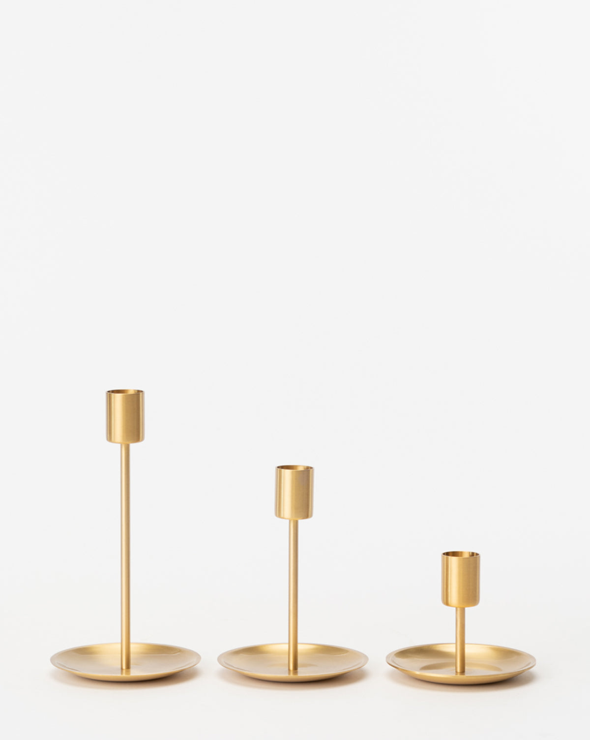 Three Brass Taper Holders, gold-colored with round bases and a sleek design in varying heights, are arranged side by side against a plain white background.
