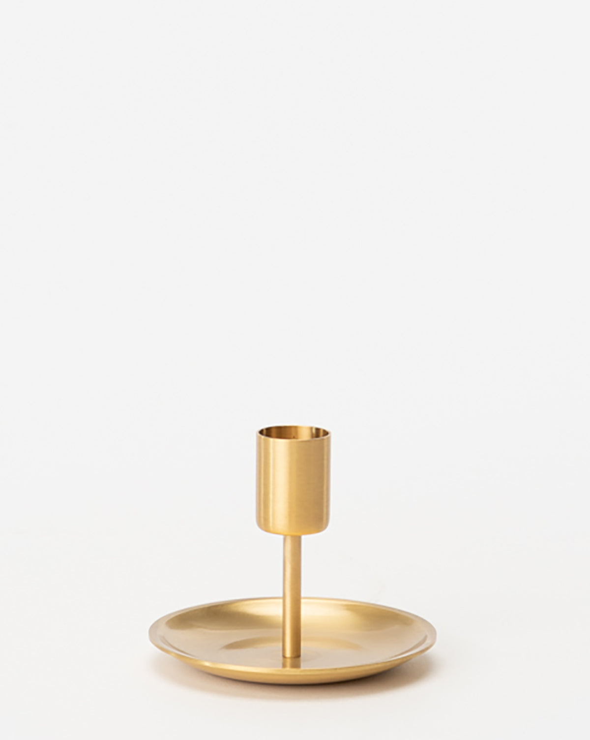 The Brass Taper Holder features a sleek, minimalist design with a round base and cylindrical holder for one candle in gold-toned metal, shown against a plain white background.