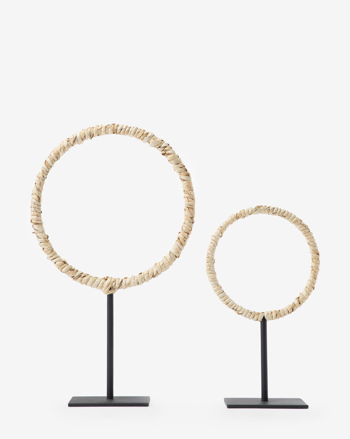 The Brayden Woven Object features two decorative circular sculptures made of small, light pieces on sleek iron stands—one large, one small—adding contemporary style to any space against a plain white background.