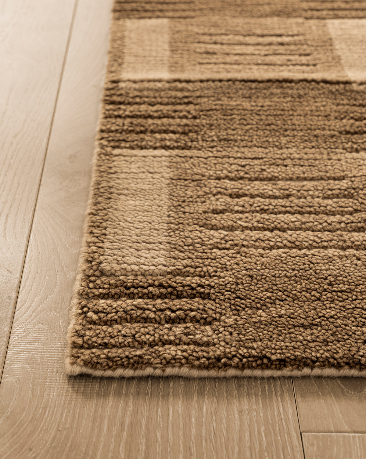 Close-up of the corner of the Brenner Hand-Knotted Wool Rug, featuring a beige and brown geometric pattern, placed on light-colored wooden flooring by McGee & Co.