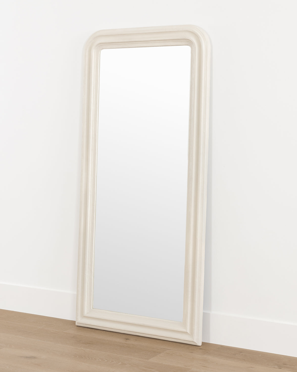 The Breslin Floor Mirror, featuring a tall, rectangular shape with a rounded top and a thick off-white plaster finish frame, leans against a white wall on a light wood floor, by McGee & Co.