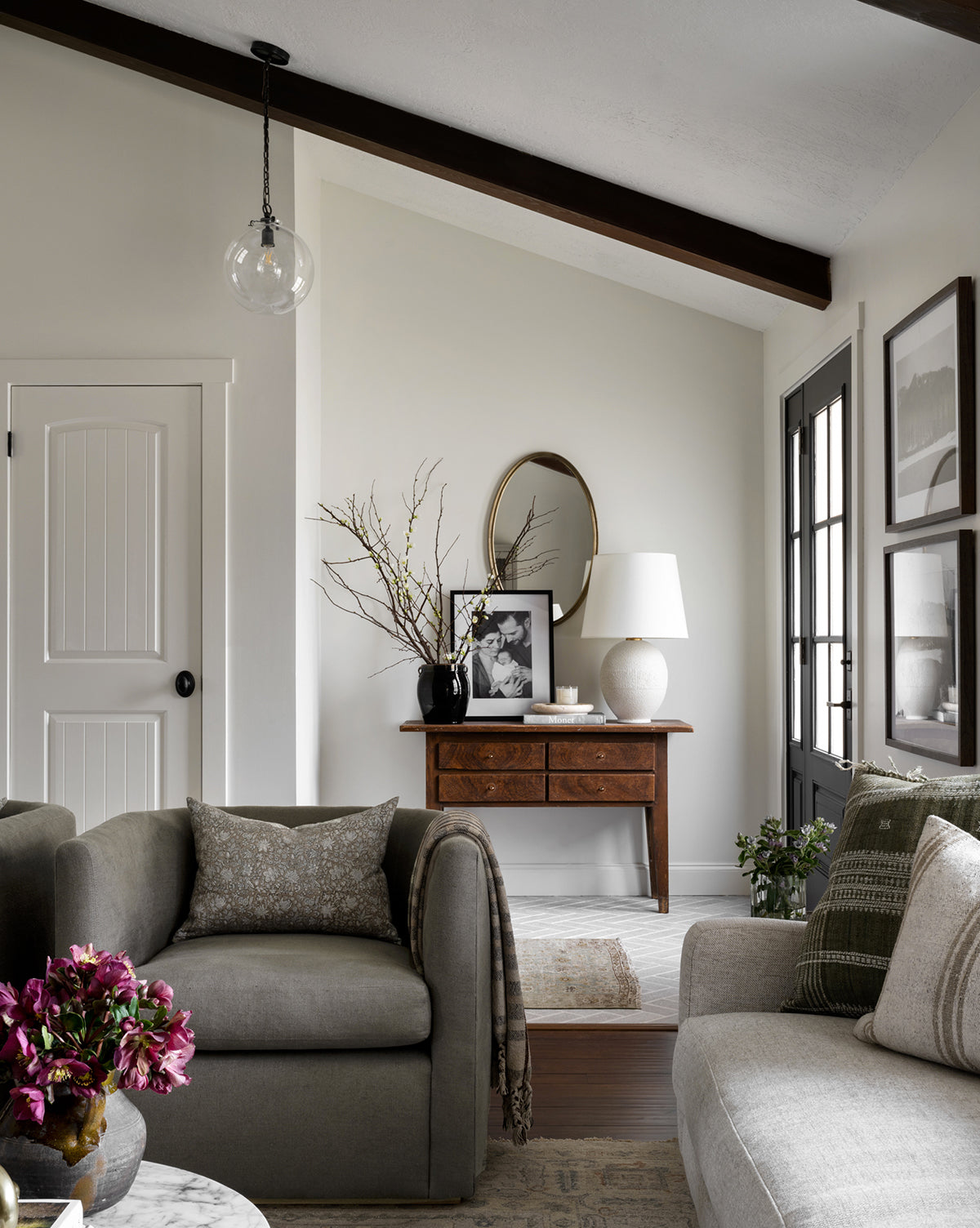 A cozy living room with neutral tones features a gray armchair, sofa, round marble coffee table with purple flowers, wooden console under a round mirror, and is illuminated by the Visual Comfort & Co. Signature Katie Globe Pendant—a setting reminiscent of the timeless elegance found at McGee & Co.