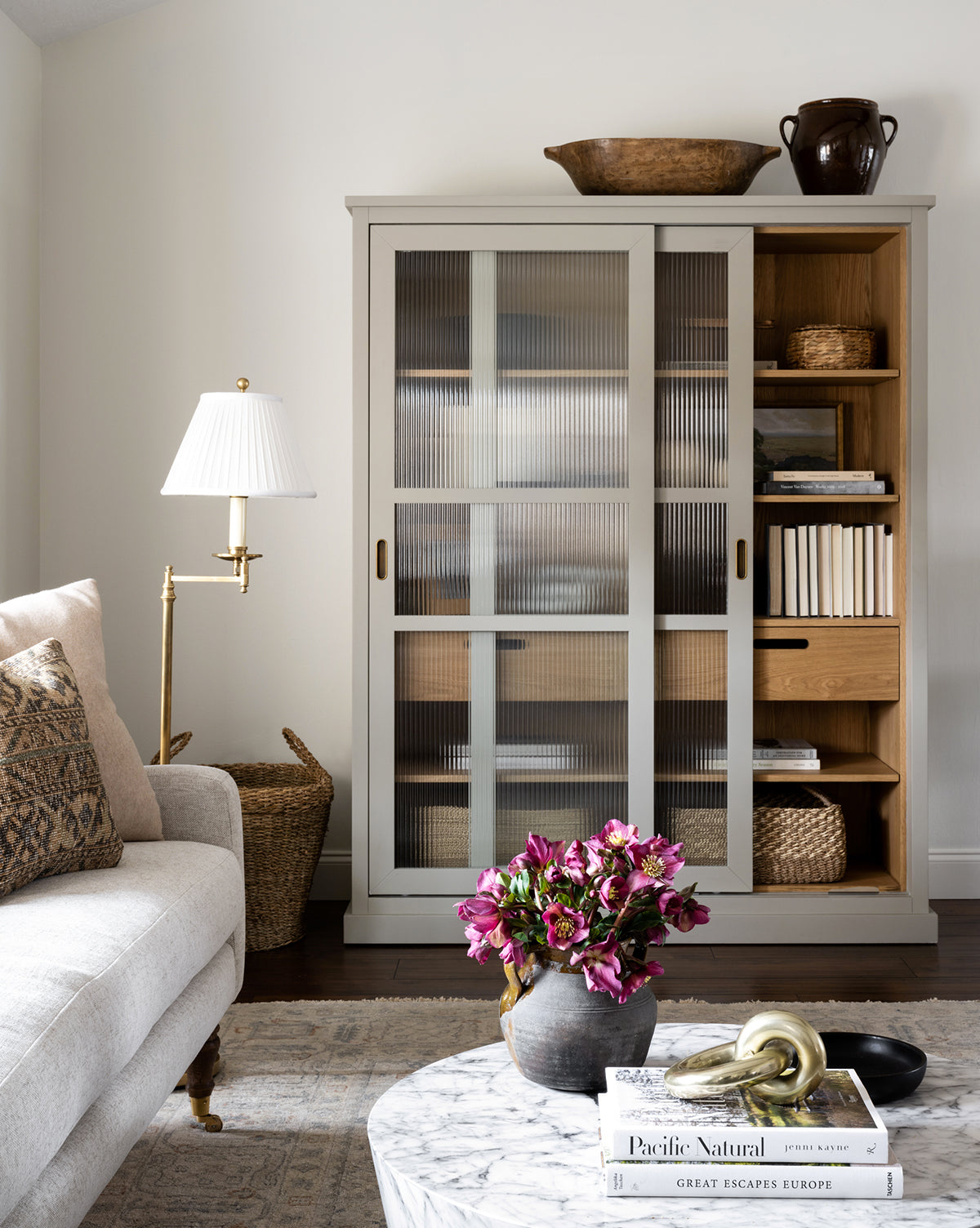A cozy living room features a beige sofa, a marble coffee table with books and flowers, and the Phillips Cabinet with reeded glass doors and antique brass handles displaying books, baskets, and ceramics. A floor lamp and woven basket sit nearby, by McGee & Co.