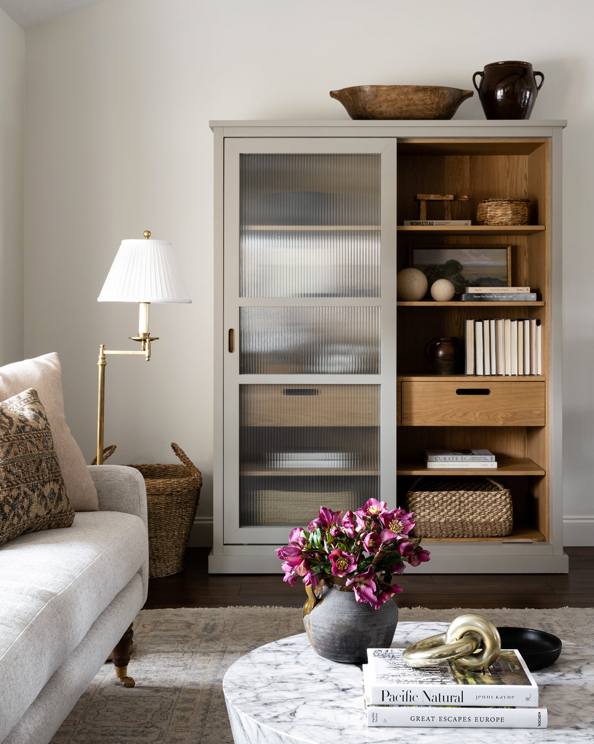 A cozy living room with a light sofa, marble coffee table with flowers, lamp, and cabinet with glass sliding doors. Shelves hold books, decorative items in warm wood and neutrals, plus the Cordova Basket for stylish storage.