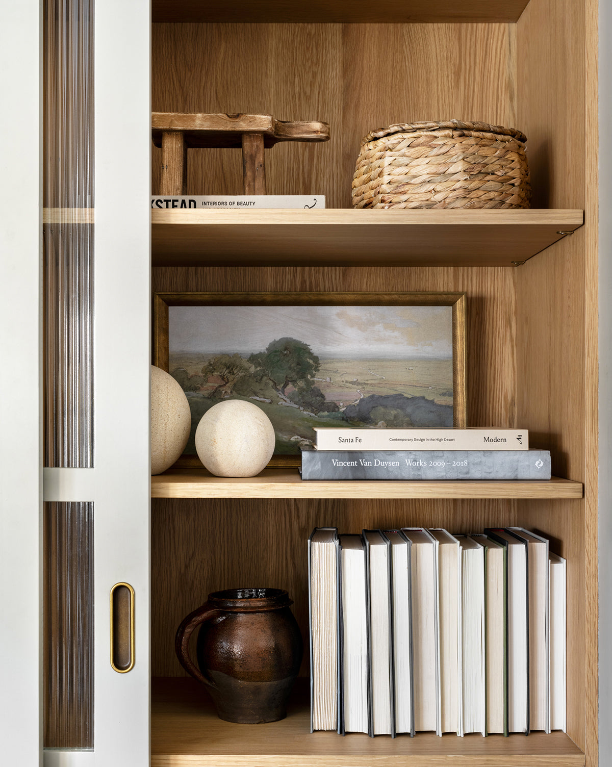 Backroads, a wooden bookshelf with a sliding glass door, showcases woven baskets, wooden decor, a framed landscape, two white orbs, a brown ceramic mug, and neatly arranged books for vintage charm.