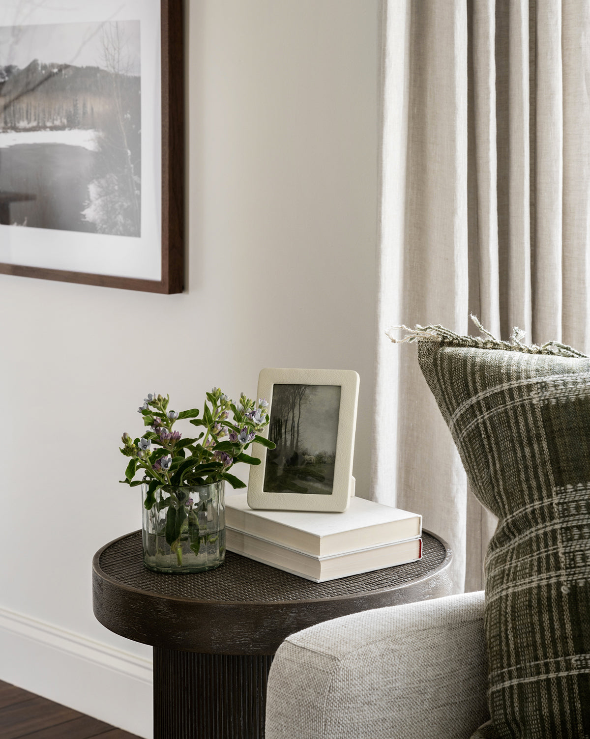 A round side table with a glass vase of purple flowers, two stacked books, and a Shagreen Picture Frame is beside a couch with a green plaid pillow. Beige curtains and a landscape photo decorate the nearby wall.