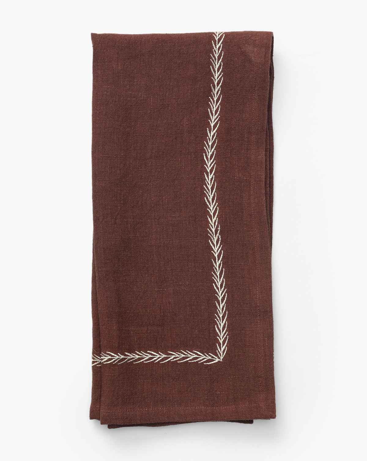 The Broadleaf Tea Towel is a folded brown linen piece featuring an embroidered leaf border at the corner, perfect for adding elegance to your kitchen, shown on a plain white background by McGee & Co.