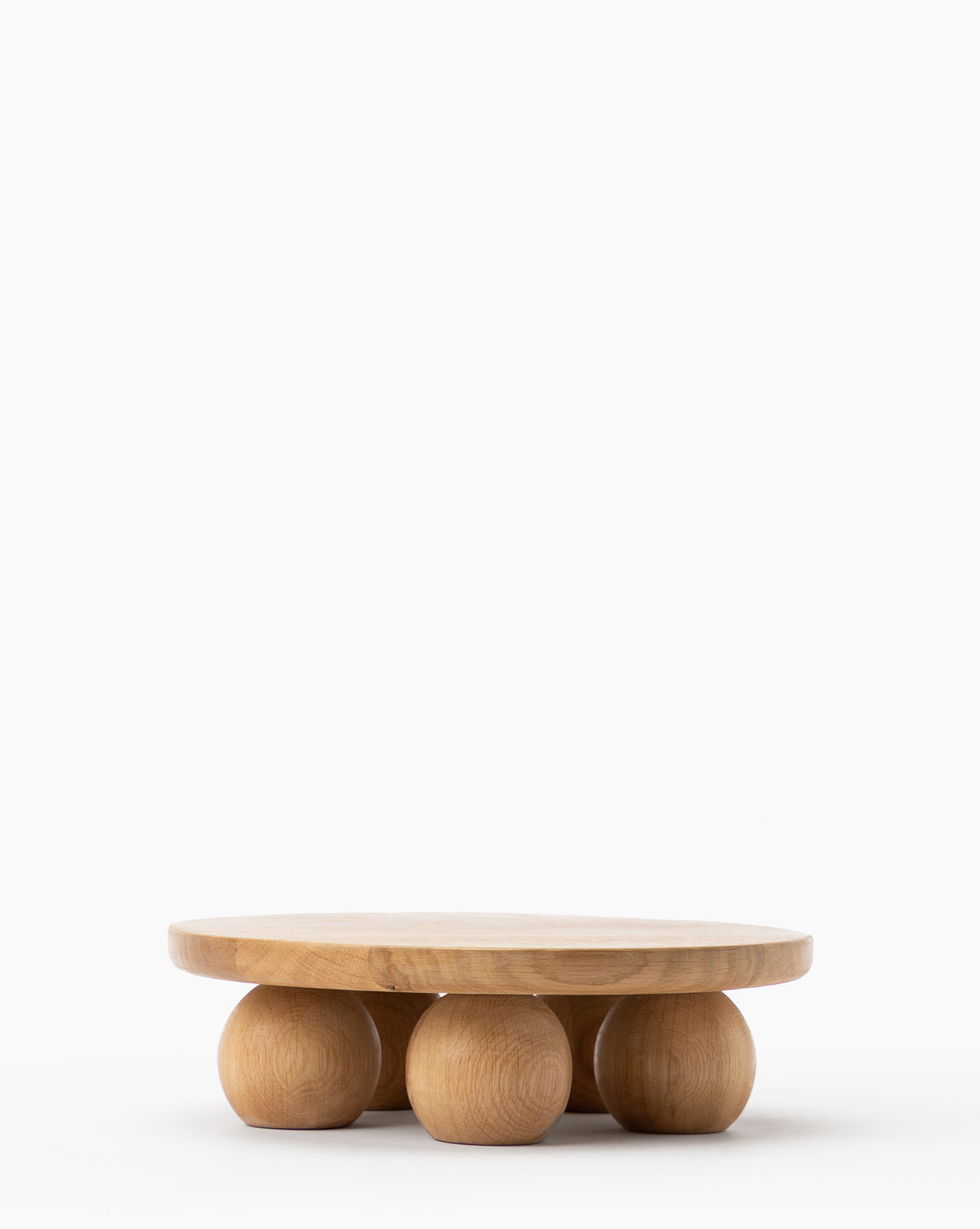 The Bronx Oak Pedestal is a low, round wooden table with a smooth top and four large spherical legs, resembling a modern cake stand, shown against a plain white background by McGee & Co.