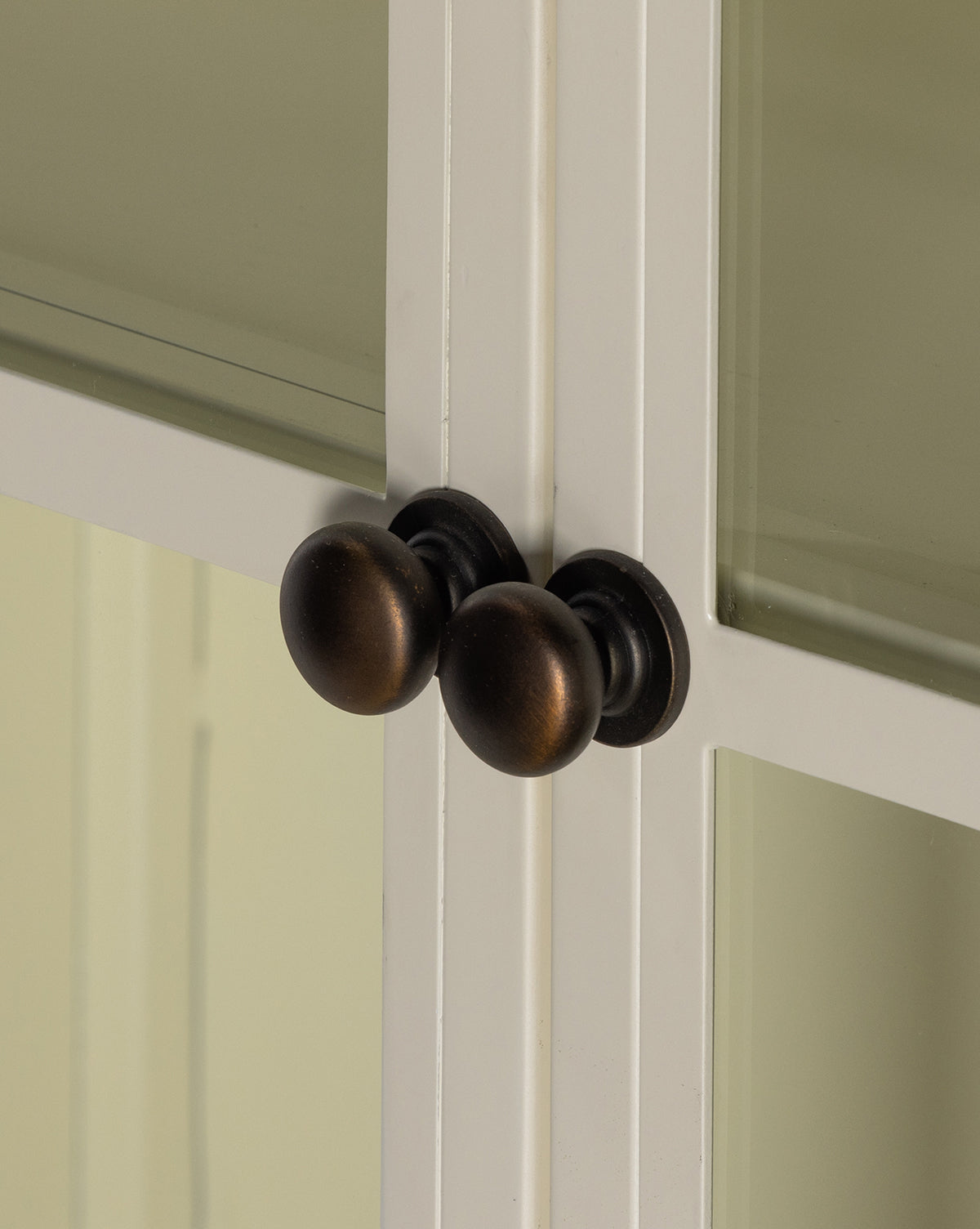 Close-up of two round, dark bronze knobs on the Brookes Cabinet’s glass doors, which are white-framed and positioned side by side where the doors meet, by McGee & Co.