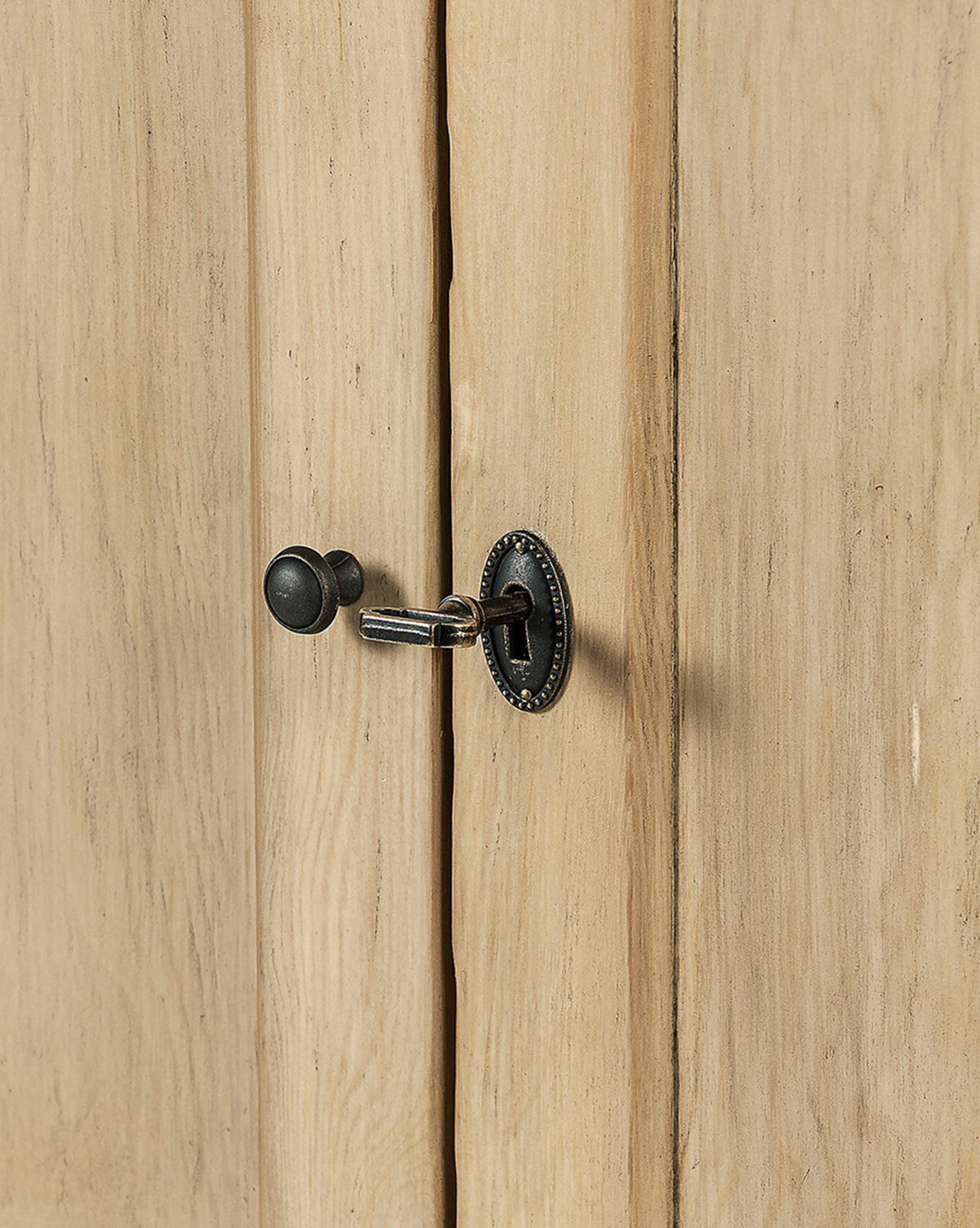 A close-up of a black metal key in an ornate keyhole on the Bryn Sideboard’s rustic pine door. The wood’s natural grain pairs with distressed copper hardware for a uniquely textured look, by McGee & Co.