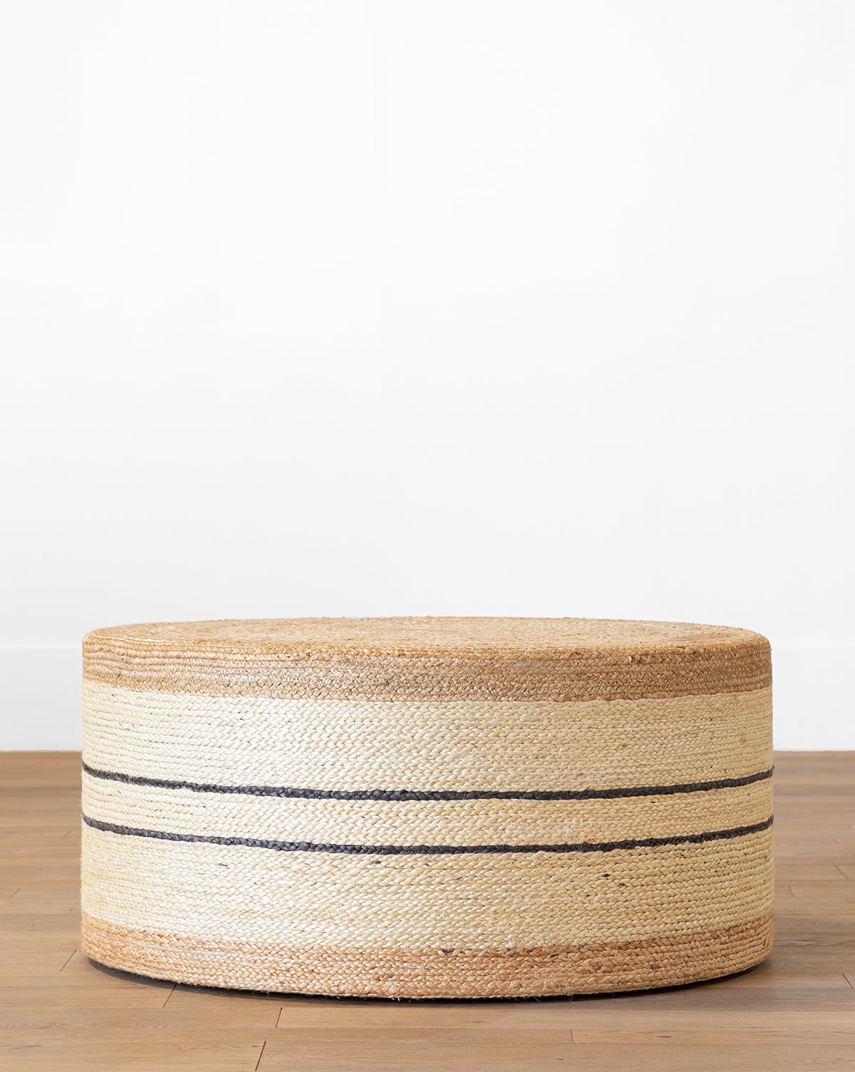 The Brynlee Woven Ottoman, round and woven with natural and tan tones plus two thin black stripes, adds a cozy accent to any seating area or bedroom. It looks great on a light wood floor against a white wall, by McGee & Co.