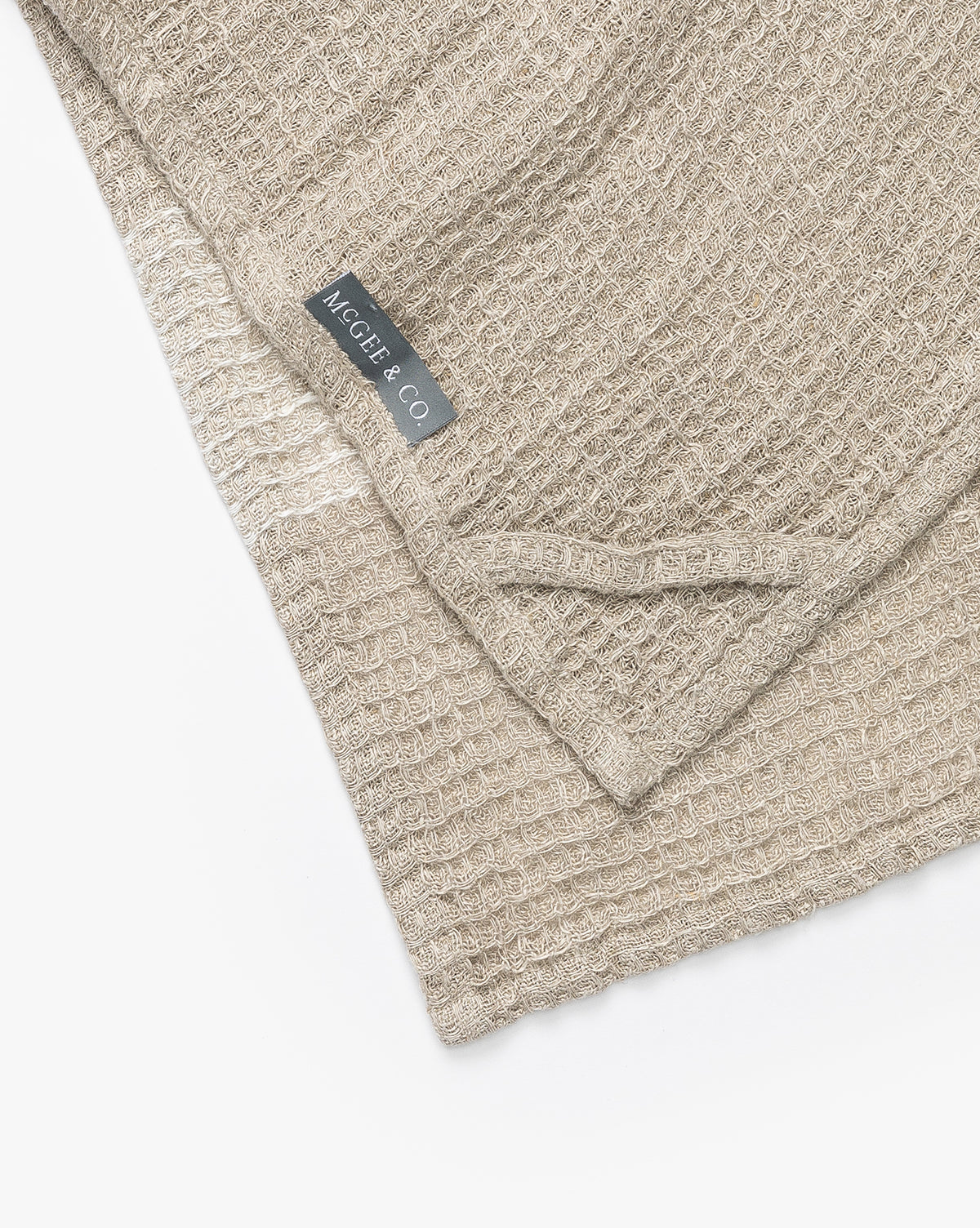The Bunton Waffle Tea Towel is a beige textured linen towel with a waffle weave, neatly folded to show a sewn-in gray NEGELLE & CO label in white text, set against a white background, by McGee & Co.