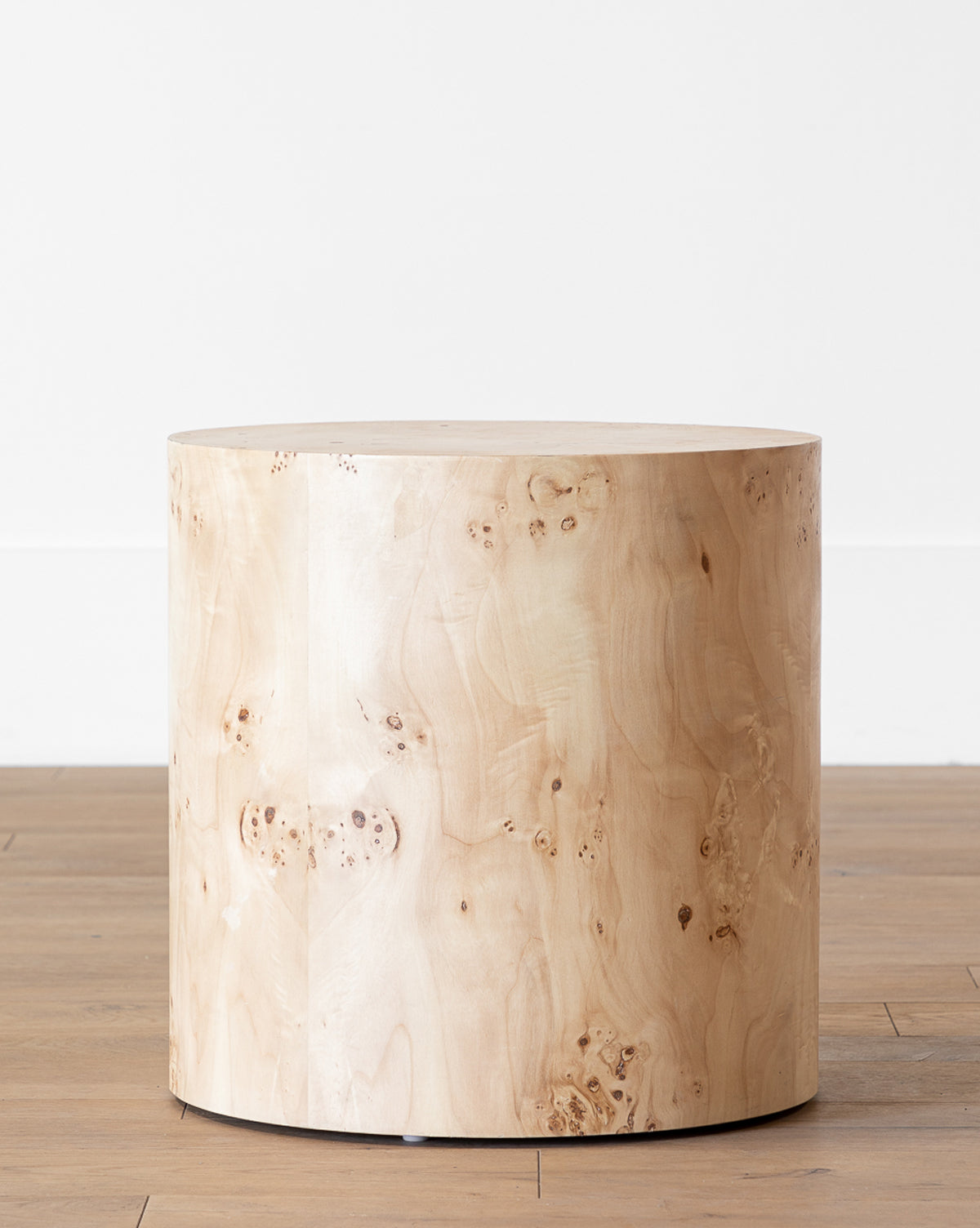 The Burl Wood Side Table is a round, modern piece with a smooth light wood finish and natural grain patterns, perfect for placing on a wooden floor against a plain white wall by McGee & Co.