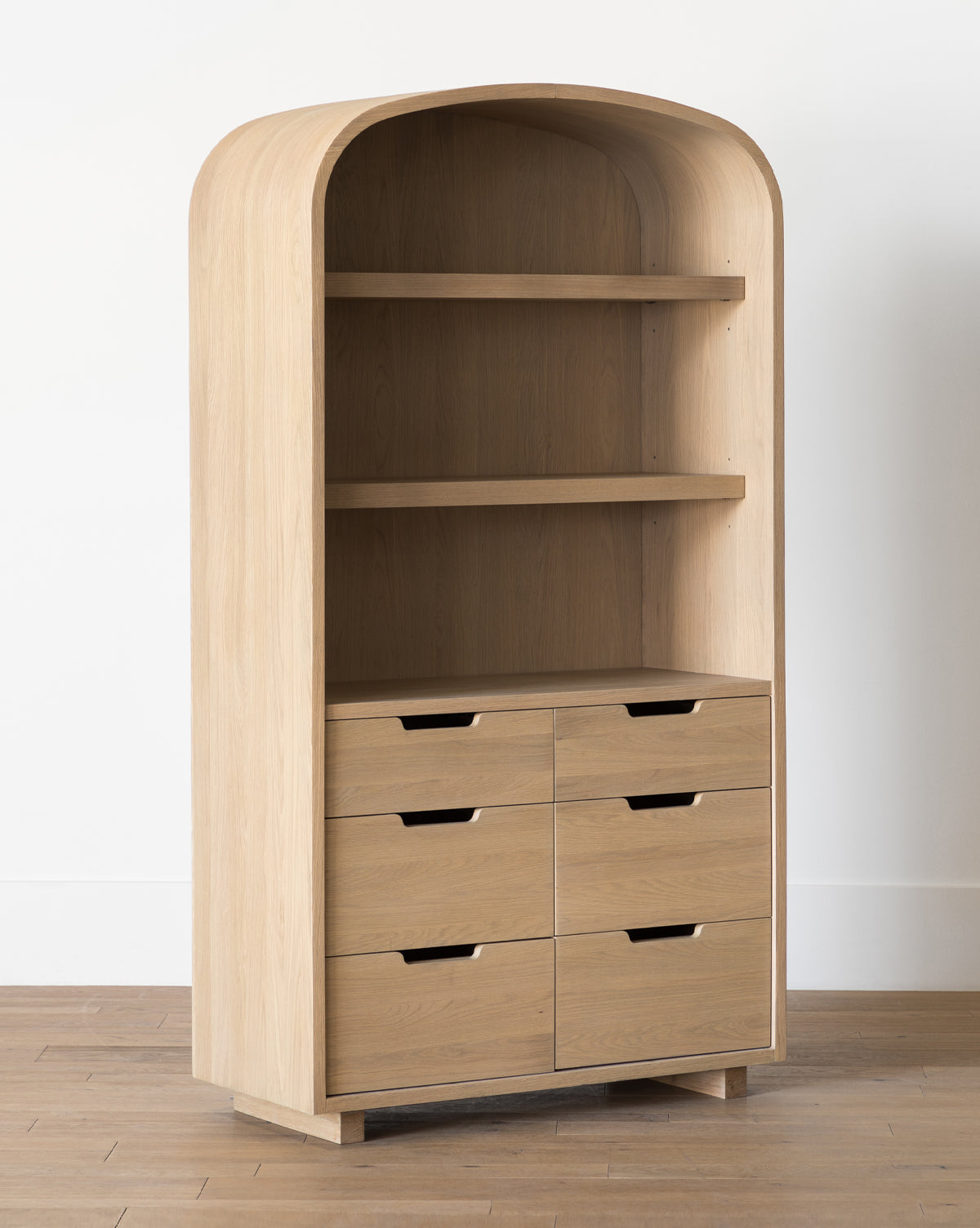 The Burnham Cabinet features light oak wood, an arched top, two open shelves, and six drawers with cut-out handles—ideal for dining room storage, by McGee & Co. It stands on a wood floor by a white wall.