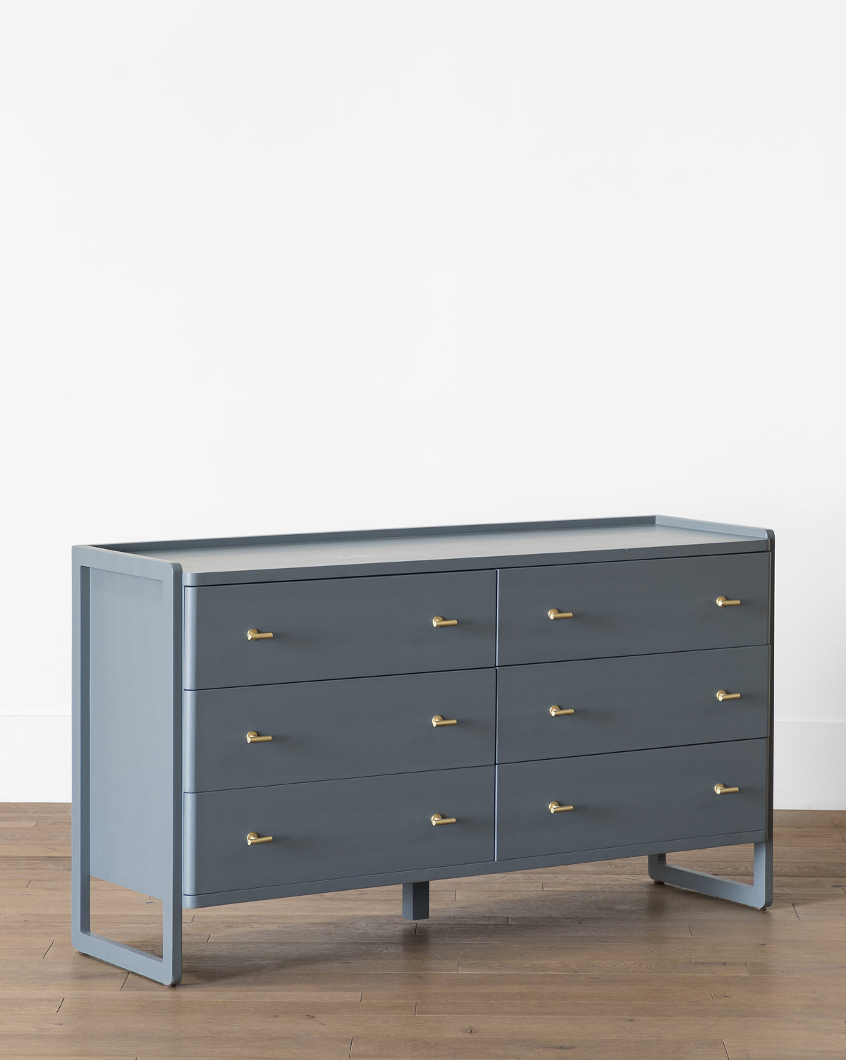 The Butler Dresser, a modern slate blue piece with six drawers and small gold handles, stands on a wood floor against a plain white wall, by McGee & Co.