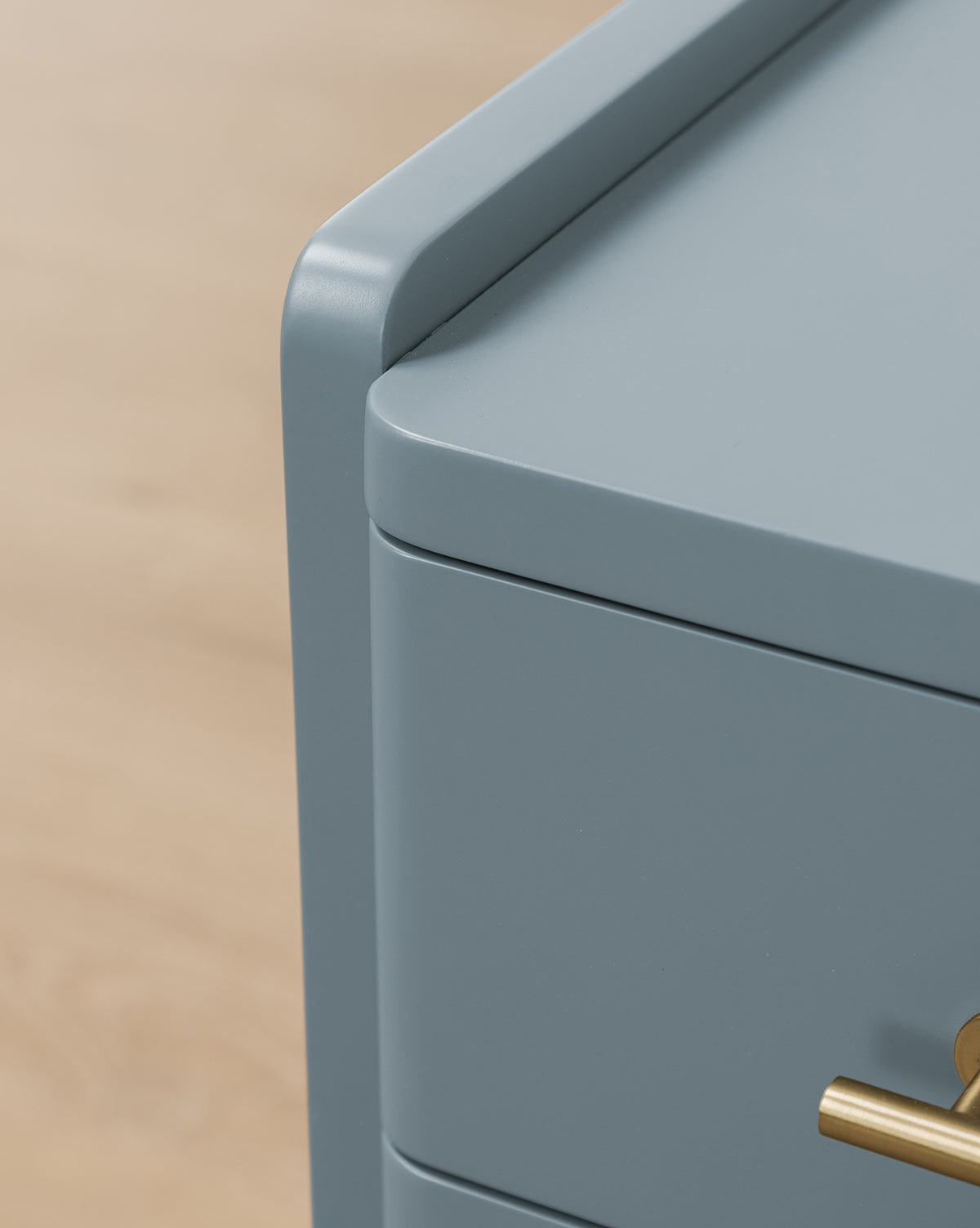 Close-up of the top corner of the Butler Nightstand in slate blue, featuring smooth, rounded edges and a brass handle, set against a light wood floor background, by McGee & Co.