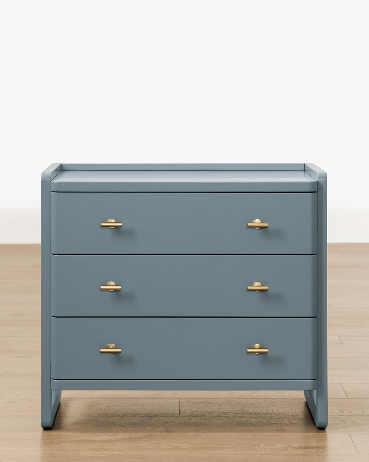 The Butler Nightstand features a modern slate blue finish, three drawers with gold handles, and stands on a light wooden floor against a plain white background, by McGee & Co.