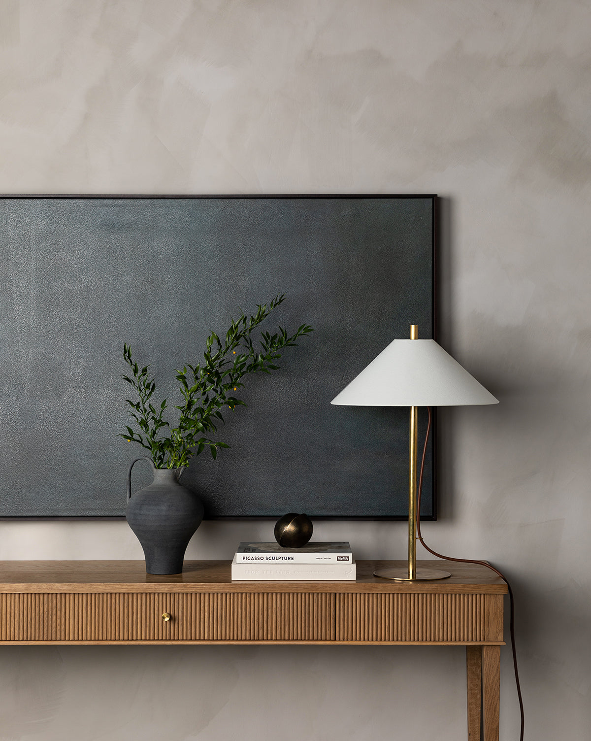 A wooden console table with a drawer holds a grey vase with green branches, a white and gold lamp, two books, and a brass sphere. The Charcoal Terracotta Jug artfully complements the large dark artwork on the textured light wall above.