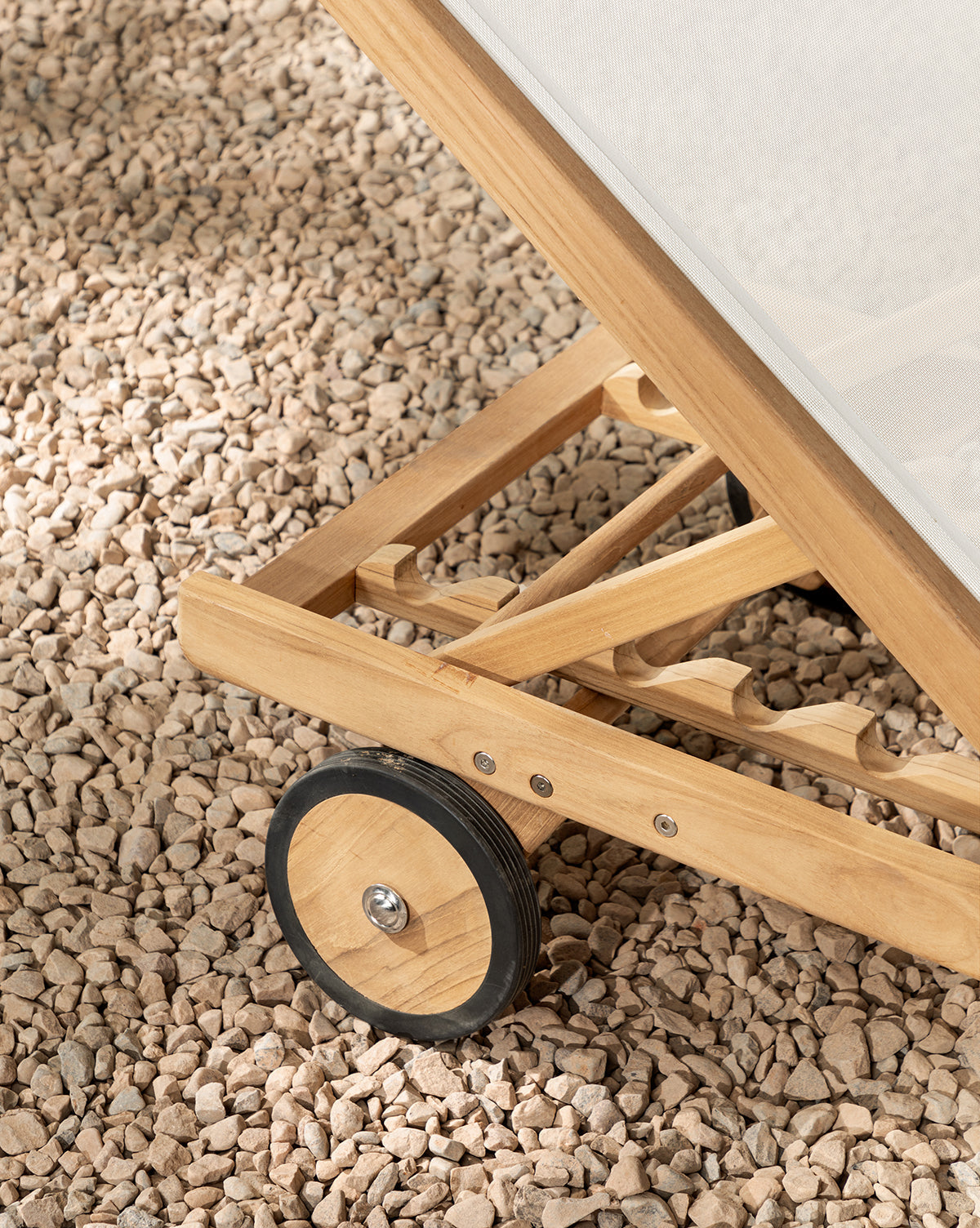 Close-up of the Cadie Outdoor Chaises teak frame and wheel on light pebbles. Featuring a weather-resistant mesh seat and an adjustable backrest, this chaise is designed for outdoor comfort by McGee & Co.