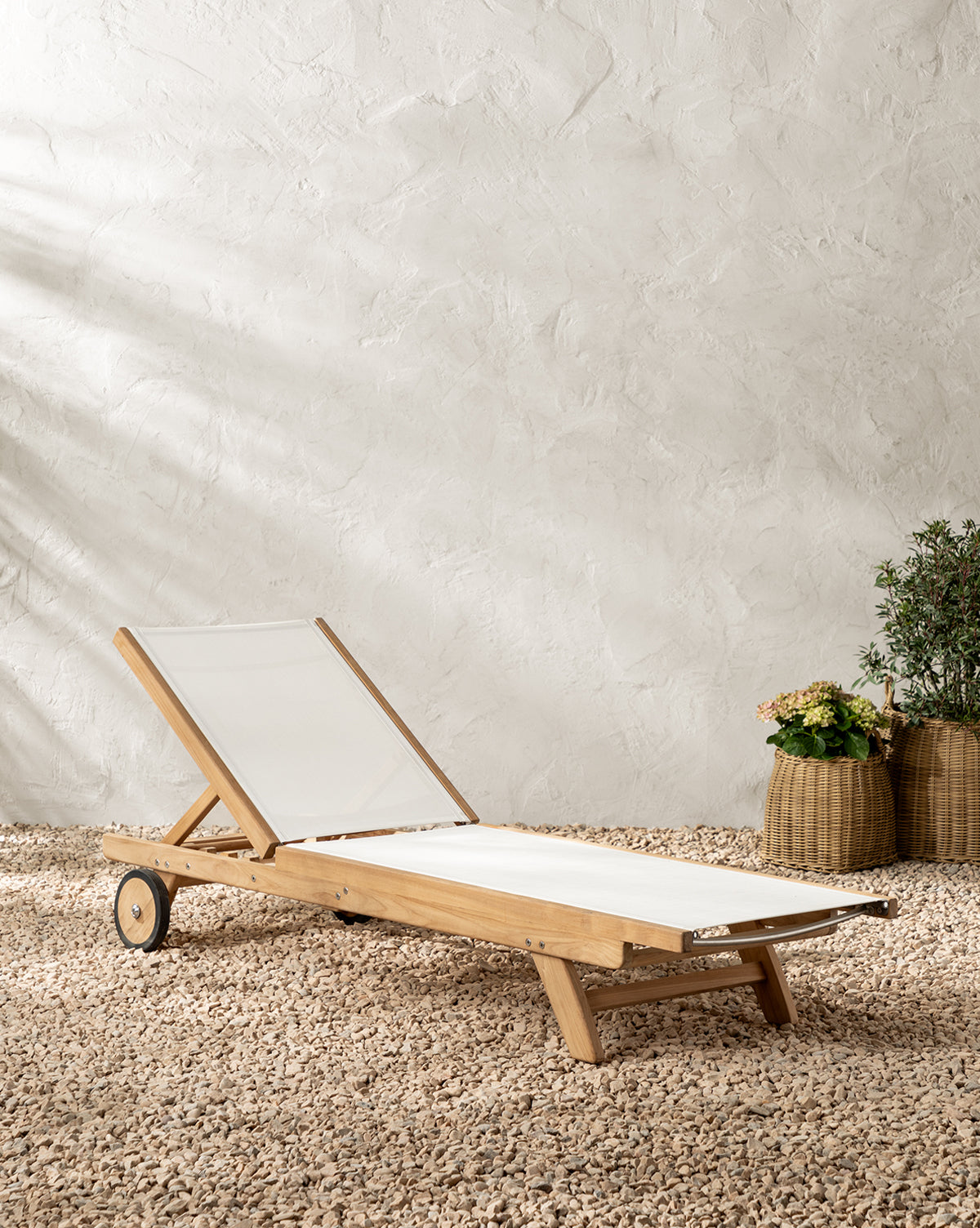 The Cadie Outdoor Chaise, featuring a teak frame and white weather-resistant cushion, rests on gravel. Nearby, two large wicker baskets with lush green plants and flowers enhance the space, set against a textured, pale wall by McGee & Co.