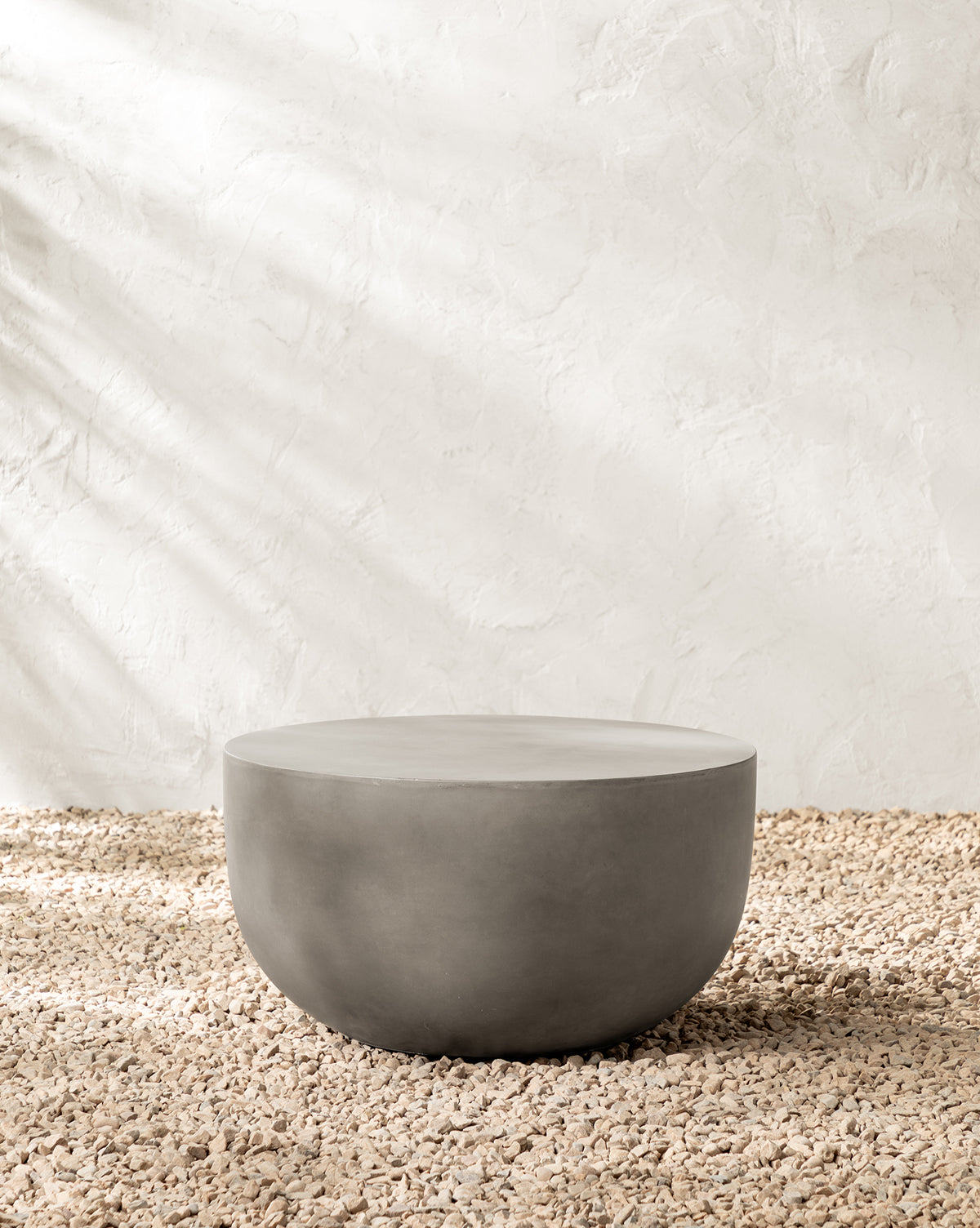 The Caine Outdoor Coffee Table, a round minimalist piece made of fiber cement, sits on a pebbled surface with a light stucco wall and natural light—perfect as modern outdoor furniture, by McGee & Co.