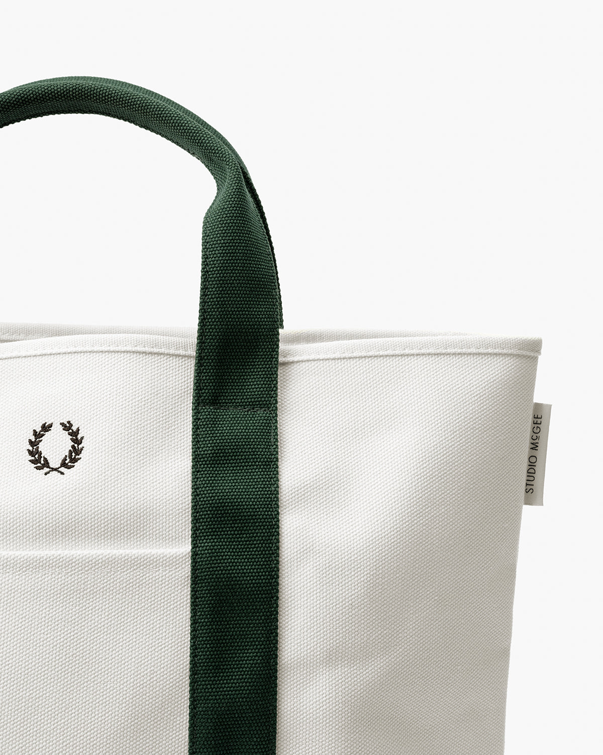 A close-up of the Canvas Laurel Tote, featuring a white canvas body, green handle, embroidered laurel wreath logo, and a small STUDIO NICHOLSON tag on the side, by McGee & Co.