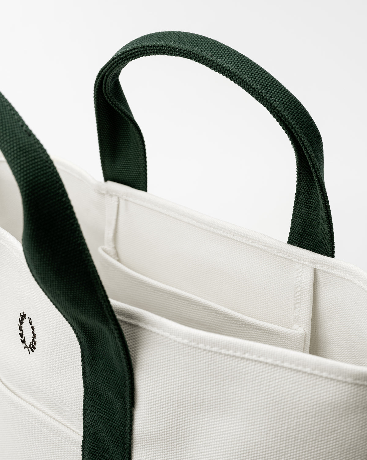 A close-up of the Canvas Laurel Tote, an everyday bag with dark green handles and trim, an embroidered wreath logo, and visible inner pockets, shown against a plain white background, by McGee & Co.
