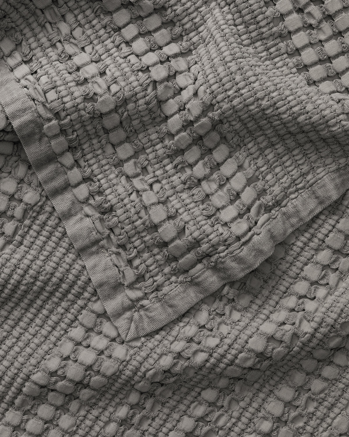A close-up of the Penedo Carnel Gray Waffle Blanket shows its solid gray, textured woven fabric with raised rectangular and circular patterns for a three-dimensional look. McGee & Co.