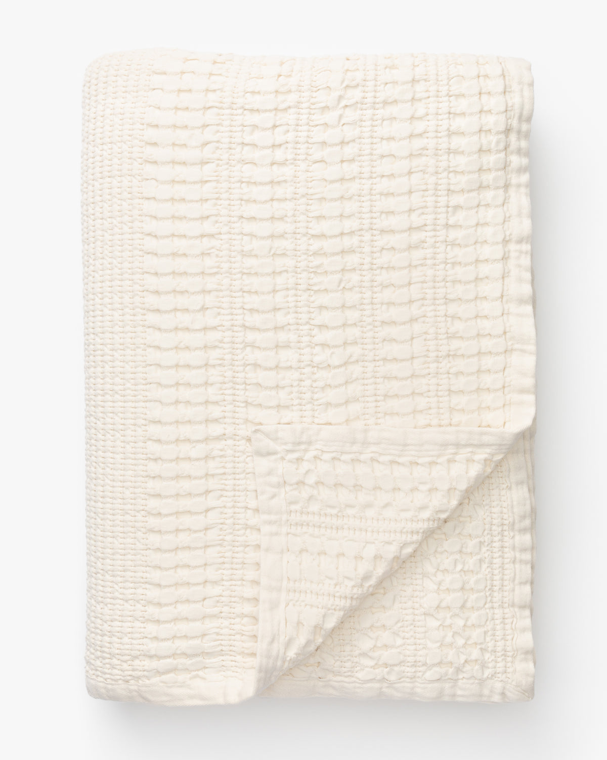 The Carnel Waffle Blanket, folded neatly against a plain white background, features a cozy textured pattern. Crafted in Portugal from 100% cotton, its cream color and inviting design add warmth to any space.