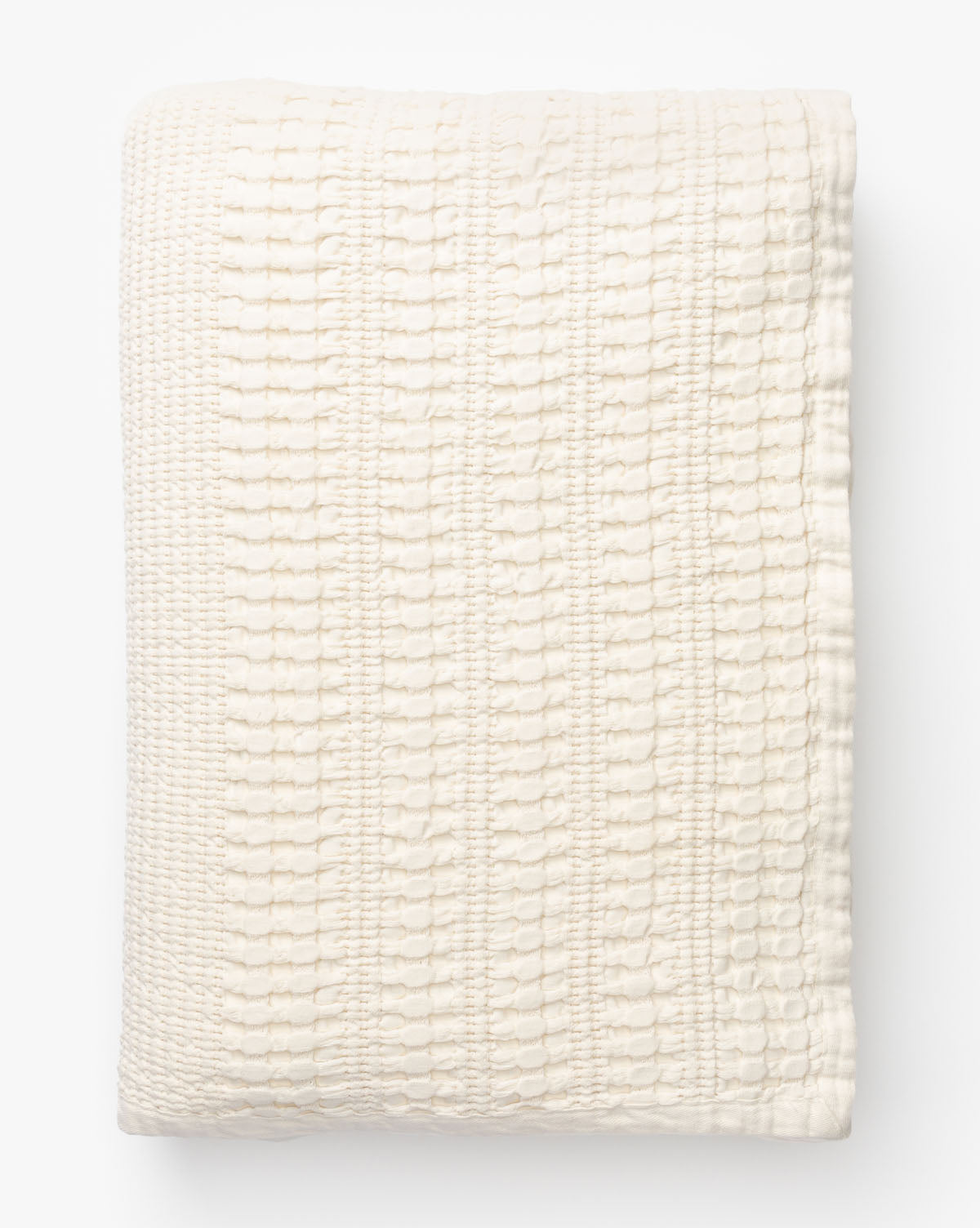 The Carnel Waffle Blanket, made from 100% cotton and featuring a textured cream-colored design, is neatly folded and stacked on a white surface against a plain white background.