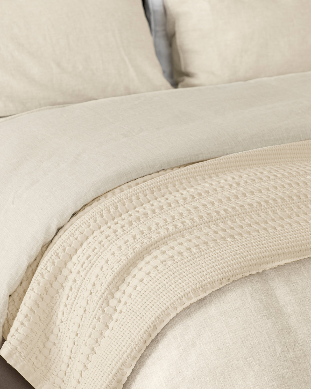 A neatly made bed features beige linen bedding and the Carnel Waffle Blanket, a textured cream layer draped at the foot. Crafted from 100% cotton, this Portugal-inspired blanket adds softness and cozy comfort to your space.