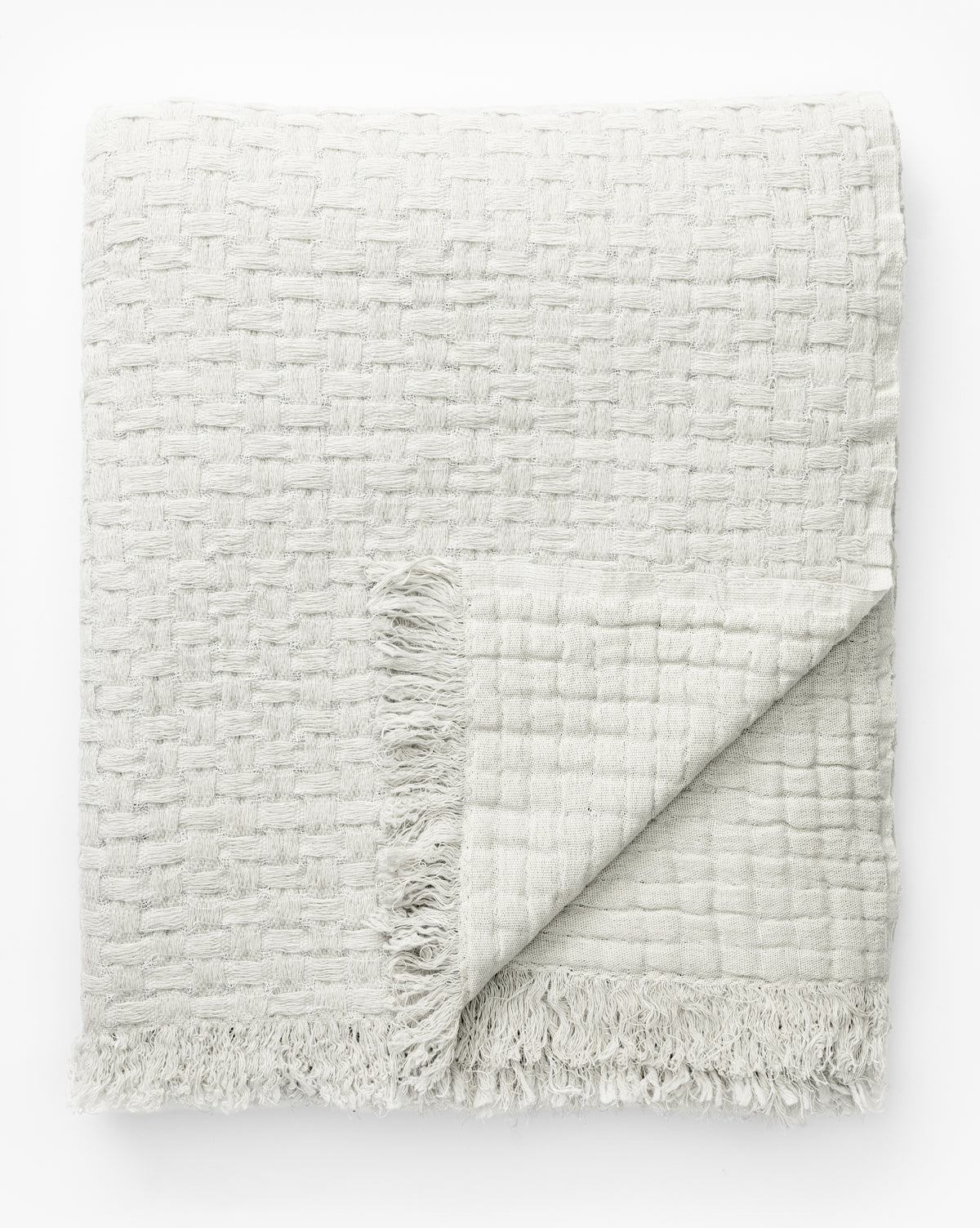 The Caroline Kids Blanket by Indigo Craft is a folded, textured off-white throw with a subtle checkered pattern and fringed edges, displayed on a white background with one corner flipped to show the reverse side. McGee & Co.