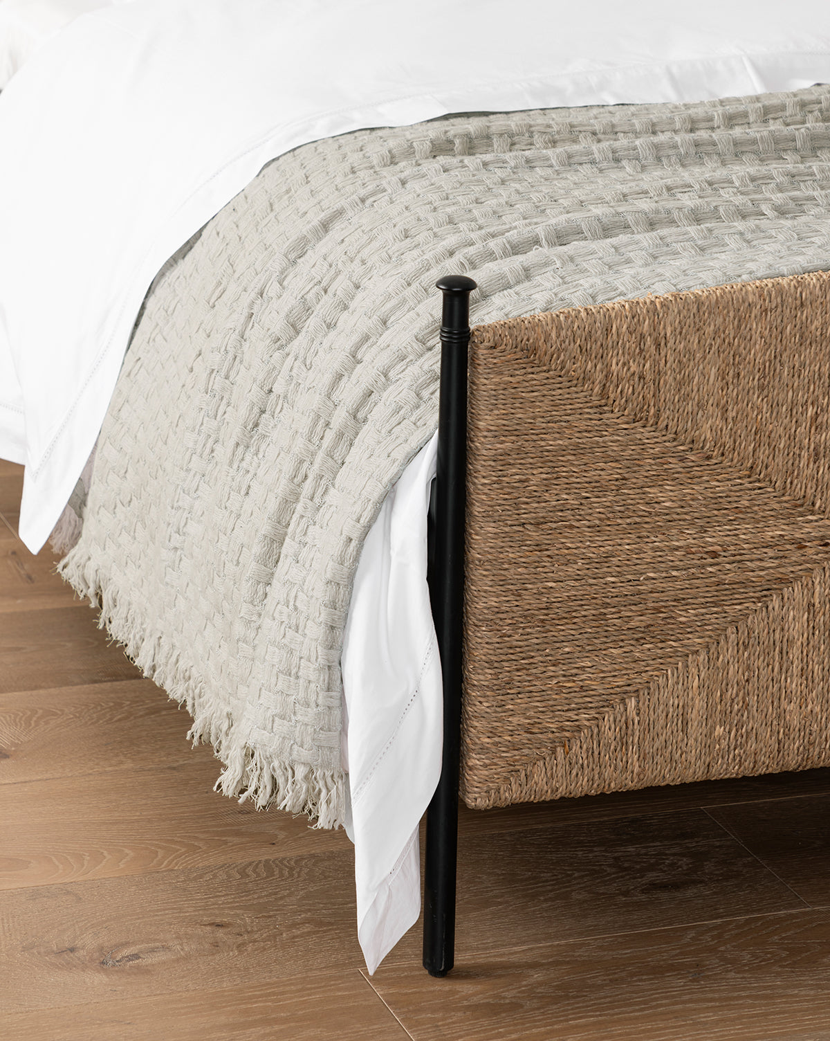 A close-up of a bed corner featuring the Caroline Kids Blanket in soft gray atop white sheets, showcased on a woven rattan bed frame with black metal posts over a light wooden floor, by McGee & Co.