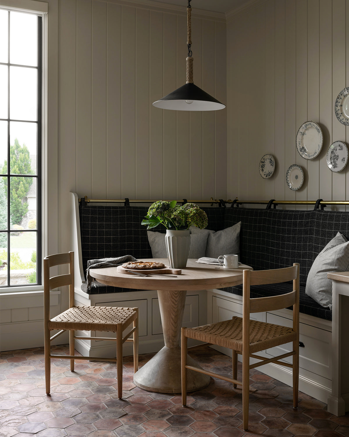 A cozy breakfast nook with Danish design features a round wooden table, Eloise Woven Chairs, a black cushioned built-in bench, a vase of green flowers, and decorative plates near a large dining room window, by McGee & Co.