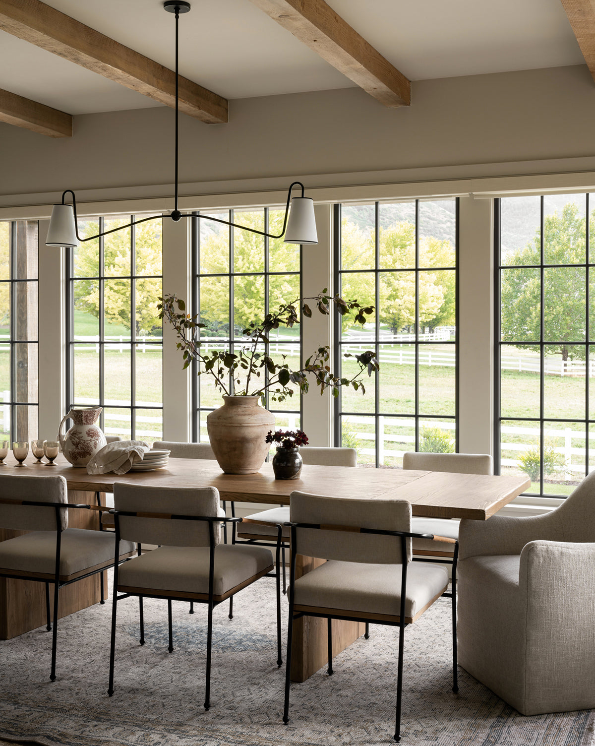 A modern dining room features a wooden table, cushioned chairs, and the Merilyn Hand-Knotted Rug. A large ceramic vase with branches sits by tall windows that overlook a green yard lined with trees and a white fence, by McGee & Co.