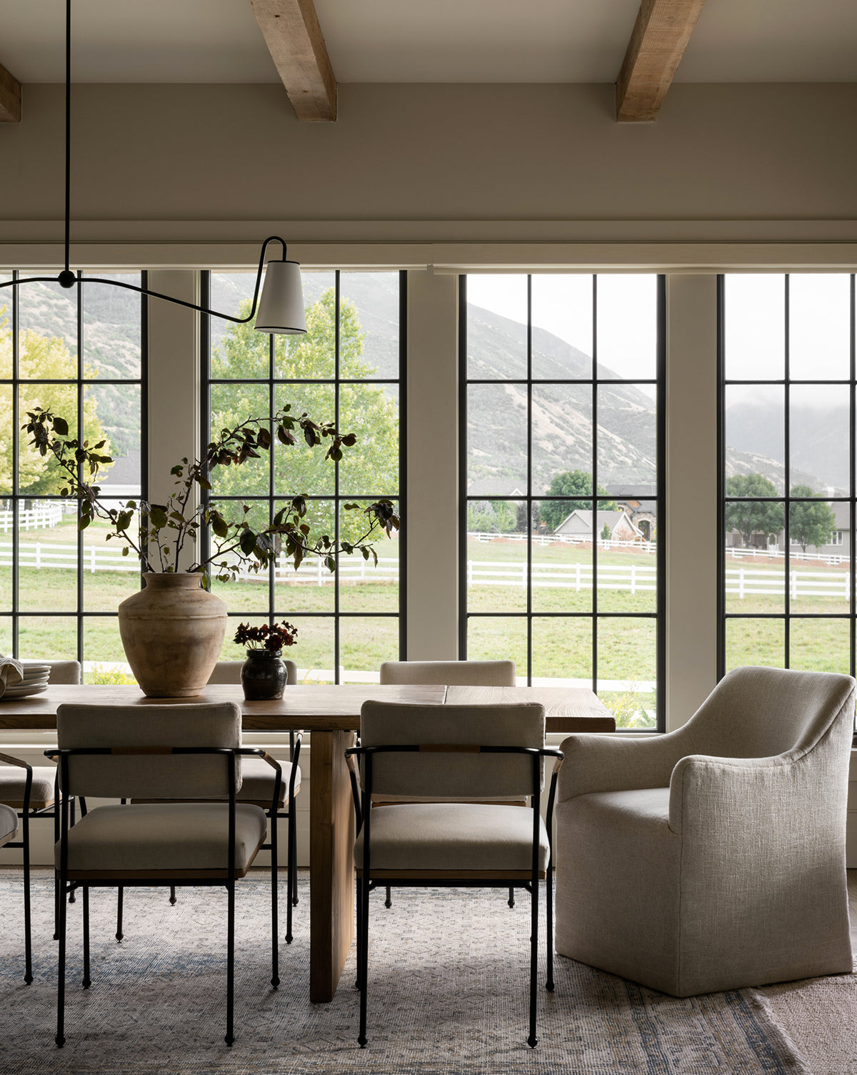 A dining room with neutral furniture, a wooden table, Elton Dining Chairs, a large potted plant centerpiece, and floor-to-ceiling windows overlooking green fields and mountains—ideal for elegant dining spaces by McGee & Co.