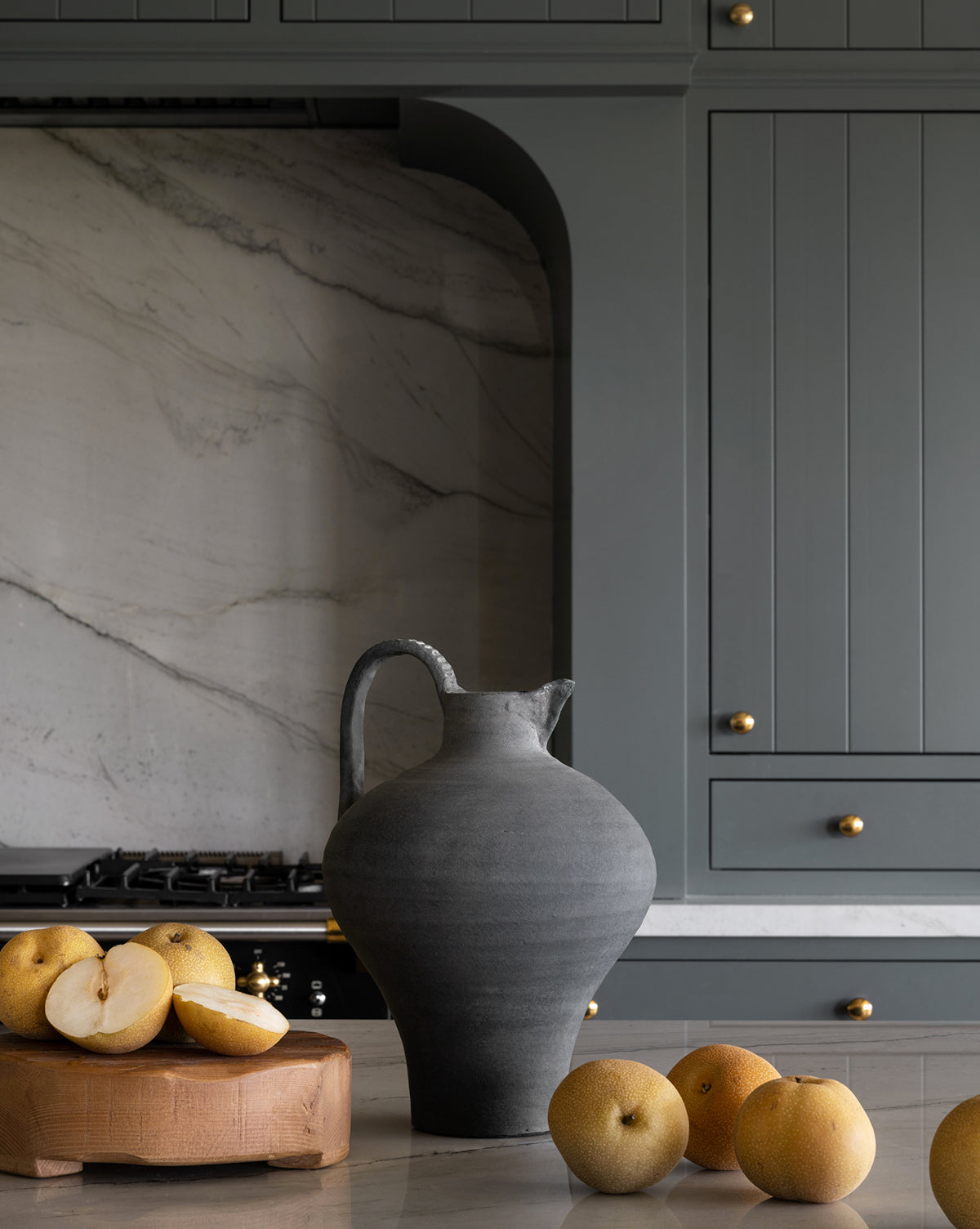 A Charcoal Terracotta Jug sits on a kitchen counter with whole and sliced Asian pears nearby, set against gray cabinets and a marble backsplash.