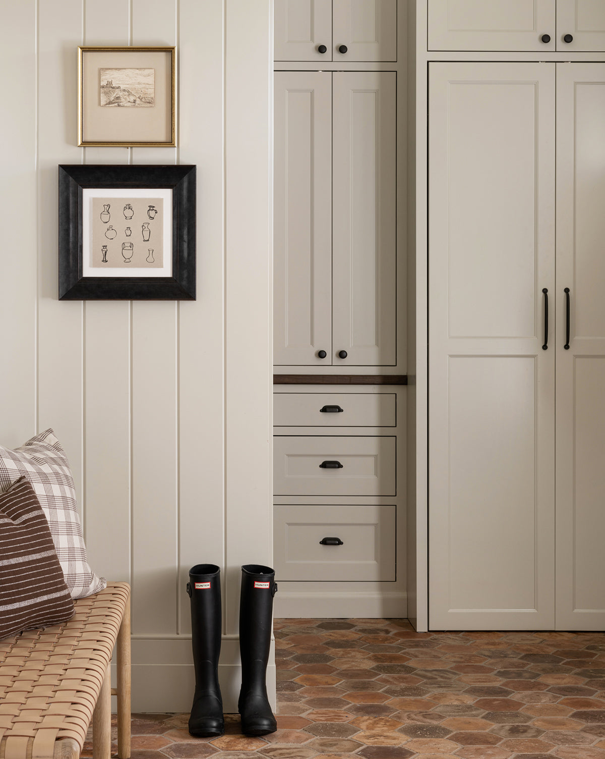 A cozy mudroom features built-in cream cabinets, a wicker bench with plaid pillows, two giclee prints in black wood frames, a pair of black rain boots on rustic tile, and a Collection of Vases displayed for added style.