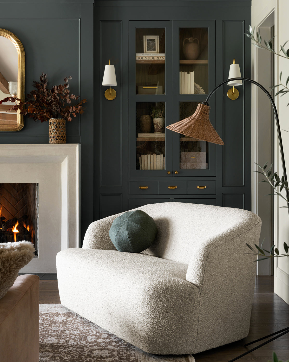 A cozy, modern living room with a light textured loveseat, green round pillow, dark built-ins, brass fixtures, a fireplace, and an arched floor lamp. Fossilized Wood Bookends and dried foliage add organic warmth to the space.