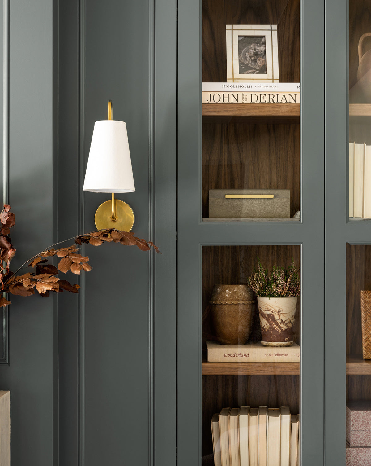 The Wonderland modern wall sconce with a white shade and gold base is mounted on a dark paneled wall beside shelves with books, art, and decor—evoking an imaginative scene reminiscent of high-fashion photography.