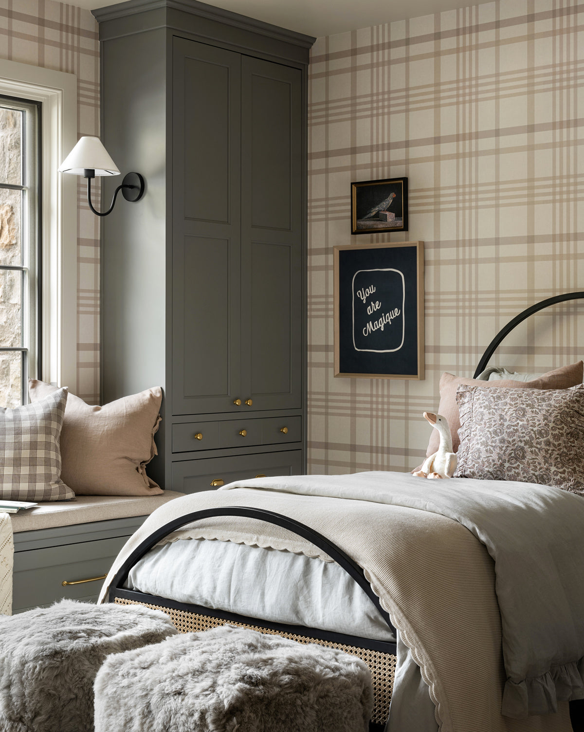 A cozy bedroom with plaid wallpaper, a bed with light bedding and furry gray stools at the foot, a window seat with pillows, built-in gray cabinets, and wall art including coastal artwork and the Blue Parrot sign.