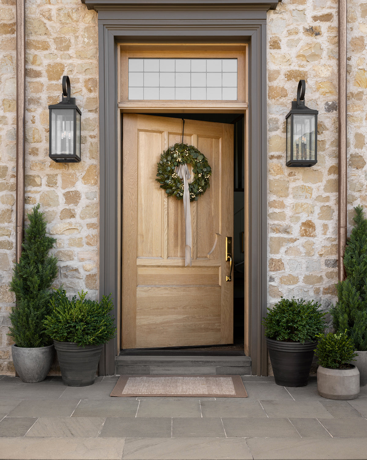 A wooden front door with a green wreath and ribbon hangs from a Metal Wreath Hanger, set in a stone wall. Black lanterns and potted evergreens flank the slightly open door, revealing a neutral doormat in the stone entryway.