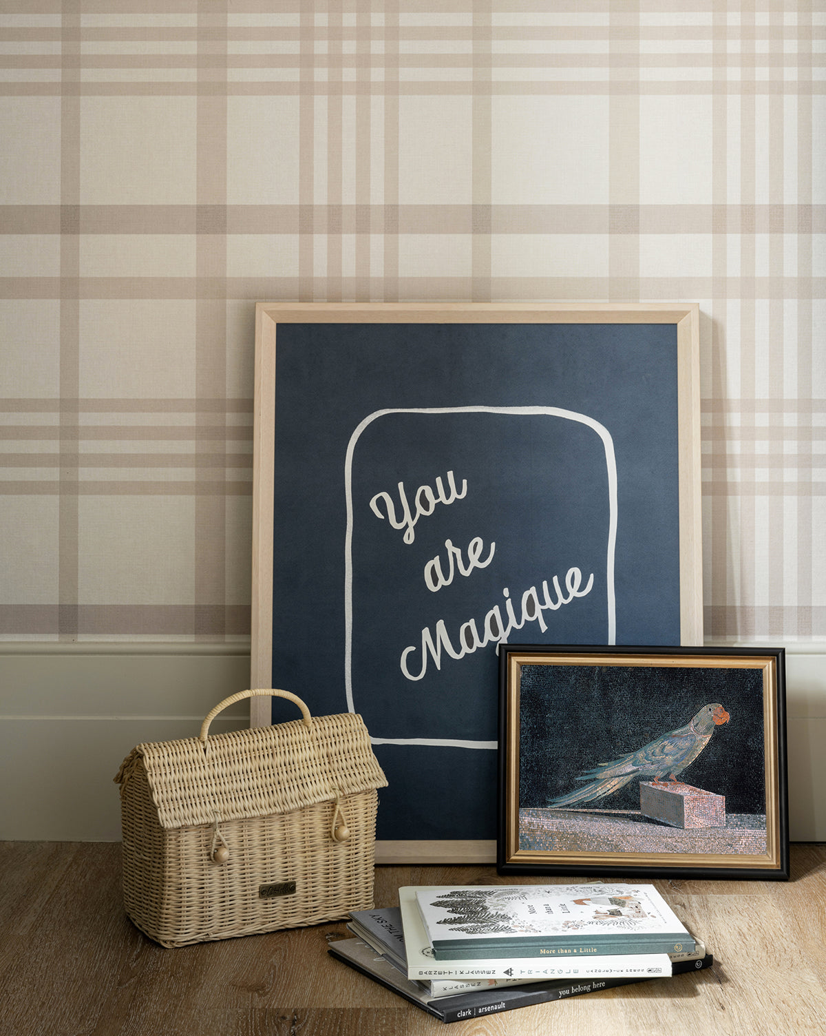 A framed giclee print reading You are Magique leans against a plaid-patterned wall. In front is the Blue Parrot framed artwork, a wicker handbag, and stacked books resting on a wooden floor.